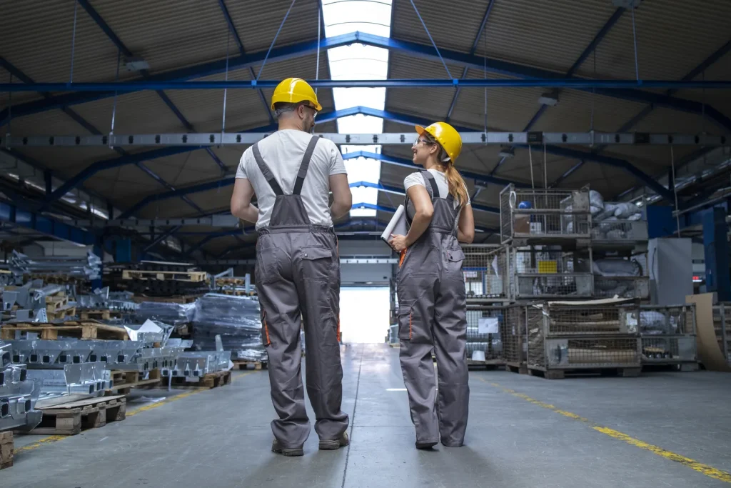 Due operai con casco e tuta da lavoro di spalle che camminano in un grande capannone industriale