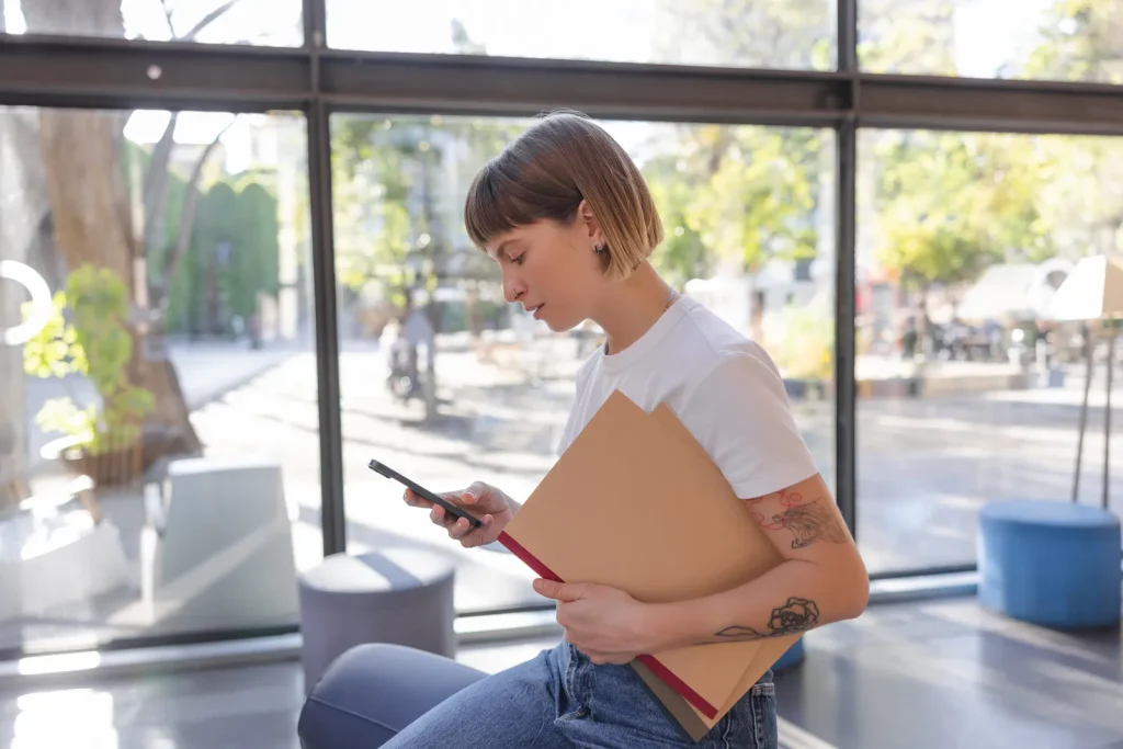 Donna con una cartellina sotto il braccio che controlla lo smartphone in un ambiente luminoso